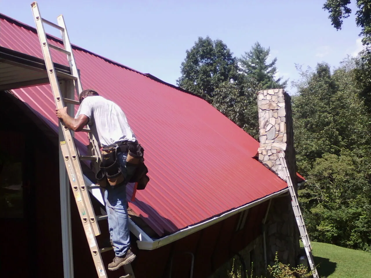 Roofer on a ladder beside a red metal roof and stone chimney — Soffit & Fascia Repair by Roofing Brothers in Hurricane Mills